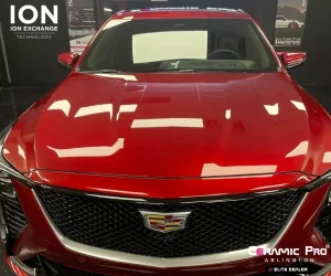 A front-view of a red luxury sedan featuring a large black mesh grille and a highly reflective hood that mirrors the overhead studio lights.