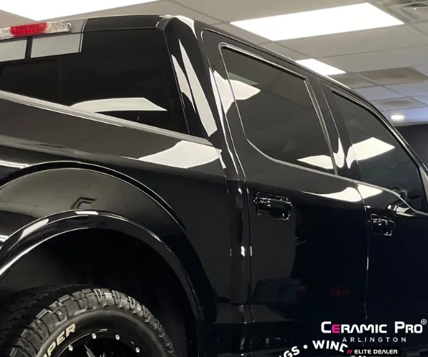 A low-angle rear view of a black pickup truck featuring a high-gloss finish and dark tinted windows inside a professional detailing shop.